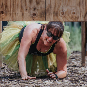 Woman wearing a tutu, climbing under an outdoor obstacle in Asskicker's Outdoor Obstacle Adventure
