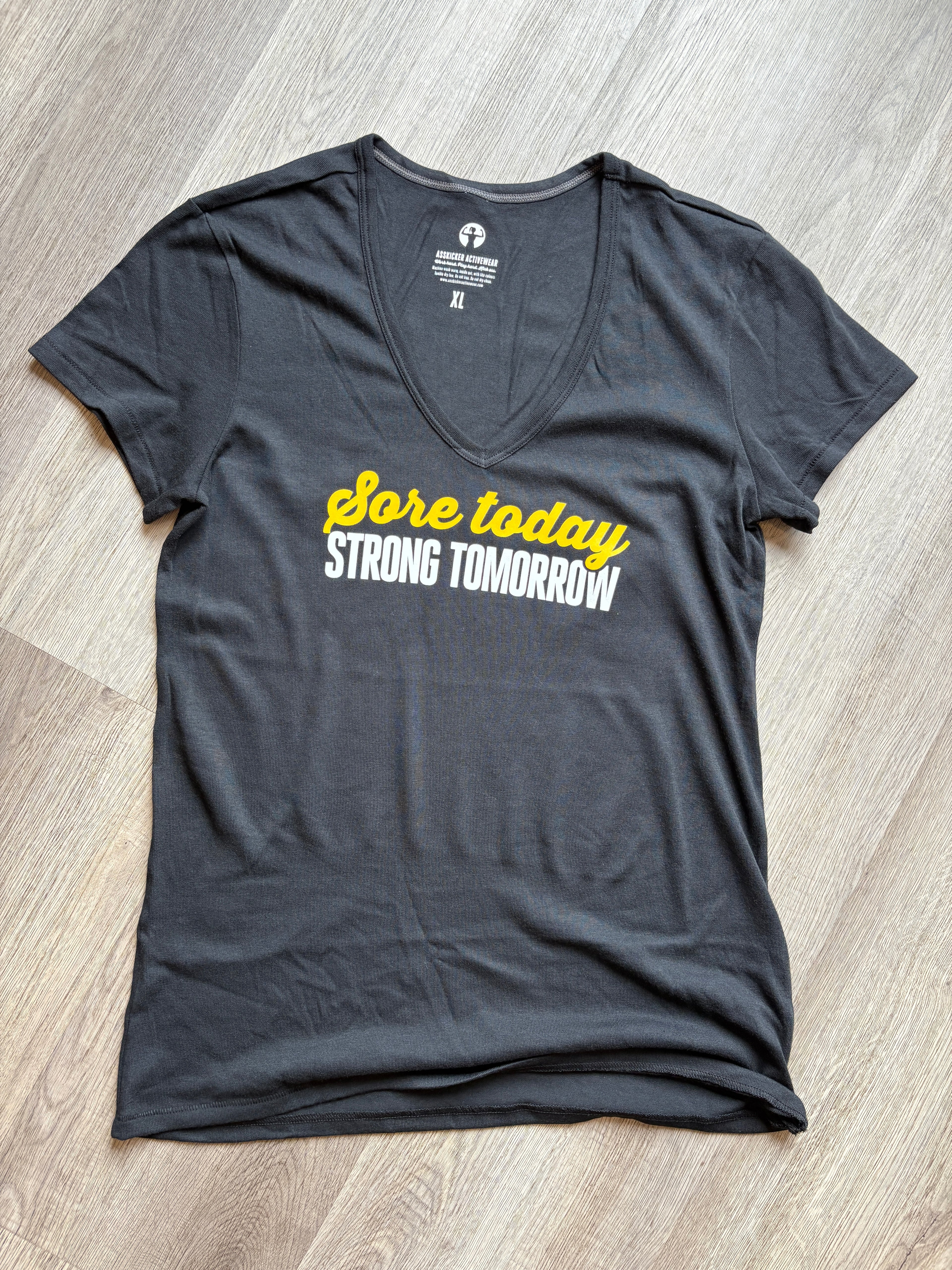 A dark gray v-neck t-shirt laid flat on a light wood floor, featuring the text 'Sore today' in yellow script and 'STRONG TOMORROW' in white block letters across the chest.