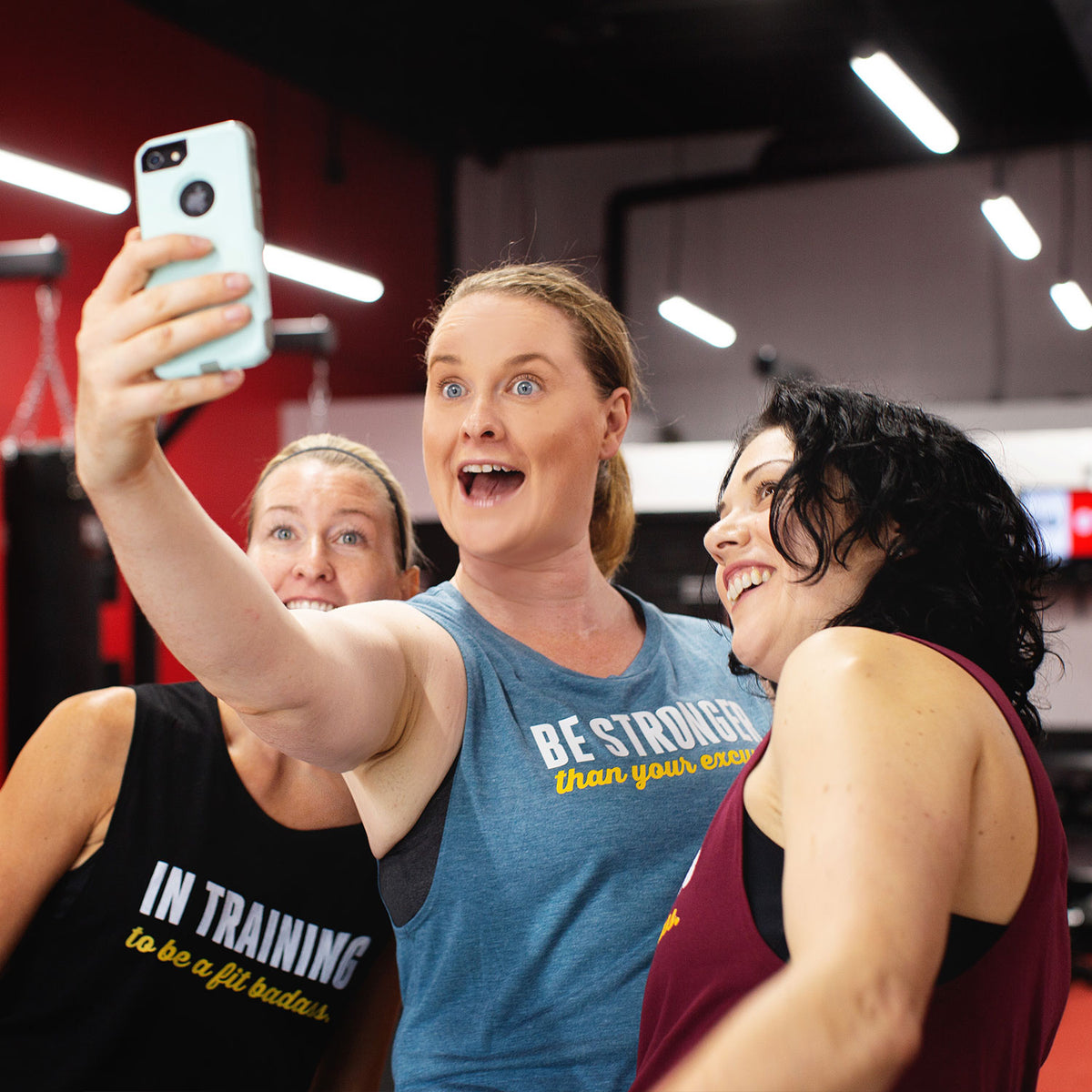 Three women taking a selfie in a gym setting, wearing flowy muscle tank tops from Asskicker Activewear, a Canadian apparel brand.