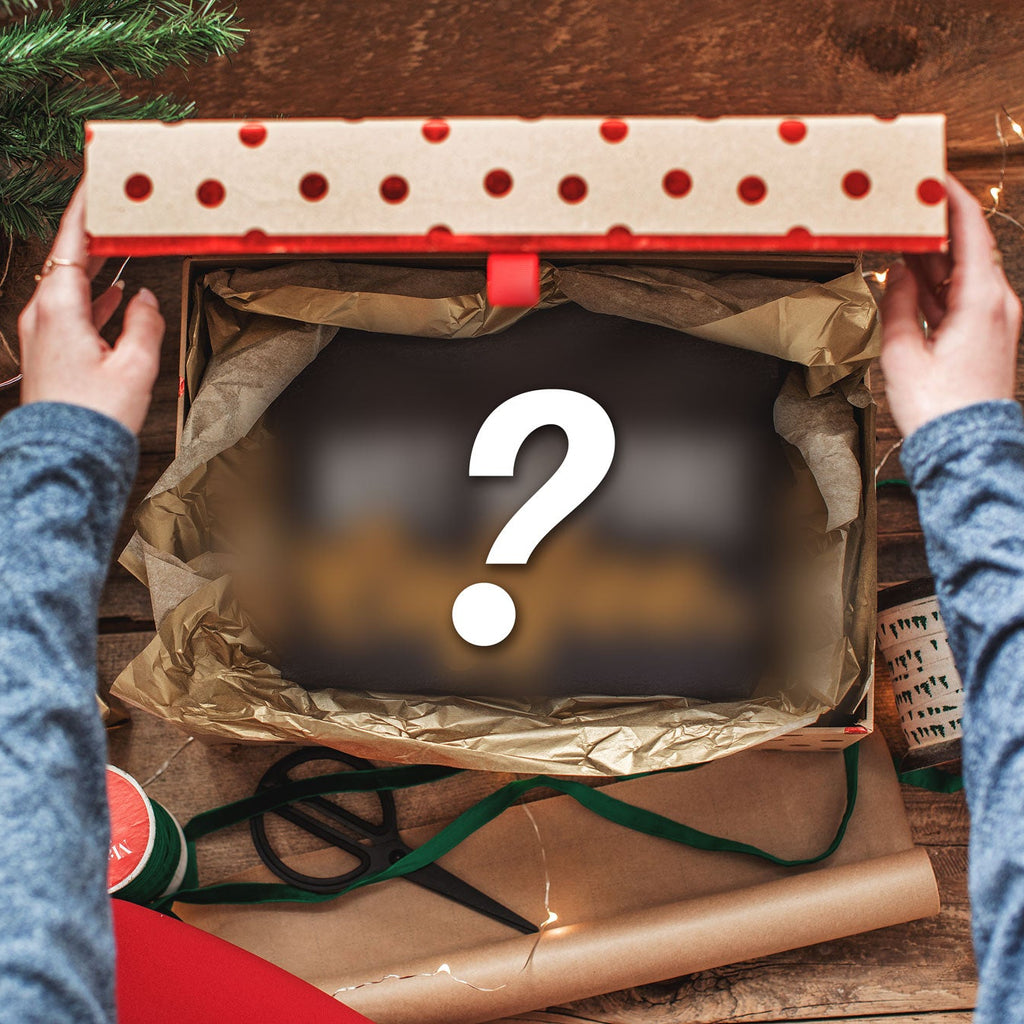 A top-down view of a person's hands holding open a gift box filled with brown tissue paper, surrounded by Christmas decorations, wrapping paper, and a large question mark graphic overlay.