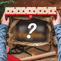 A top-down view of a person's hands holding open a gift box filled with brown tissue paper, surrounded by Christmas decorations, wrapping paper, and a large question mark graphic overlay.