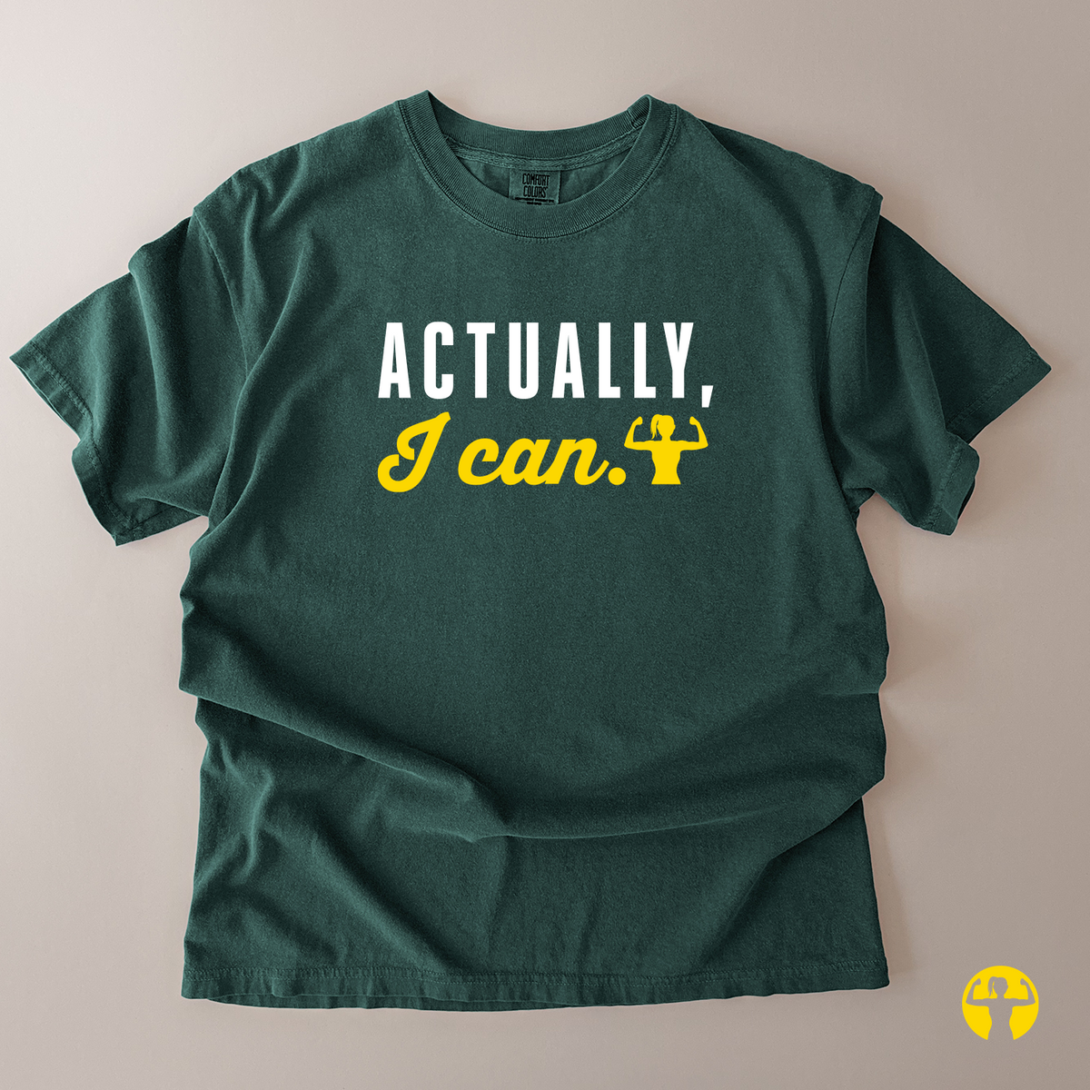 A dark green short-sleeve t-shirt laid flat on a light beige background, featuring the text 'ACTUALLY, I can.' printed on the chest in white and yellow, with a small yellow flexing arm graphic.