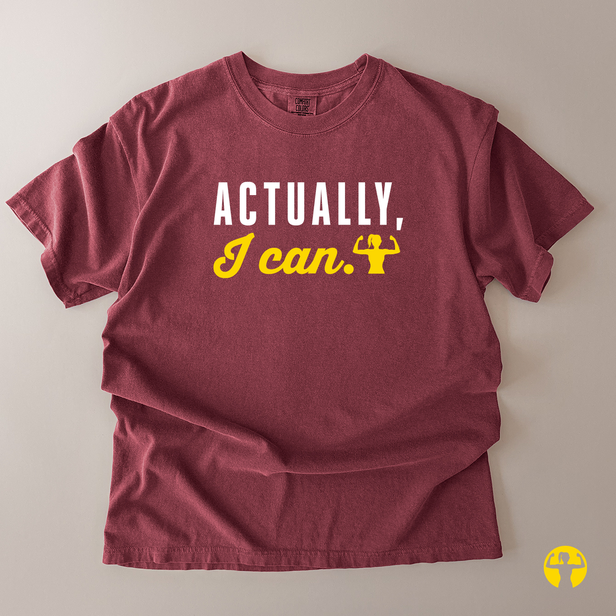 A maroon short-sleeve t-shirt with the text 'ACTUALLY, I can.' printed on the chest in white and yellow, laid flat on a light beige background.