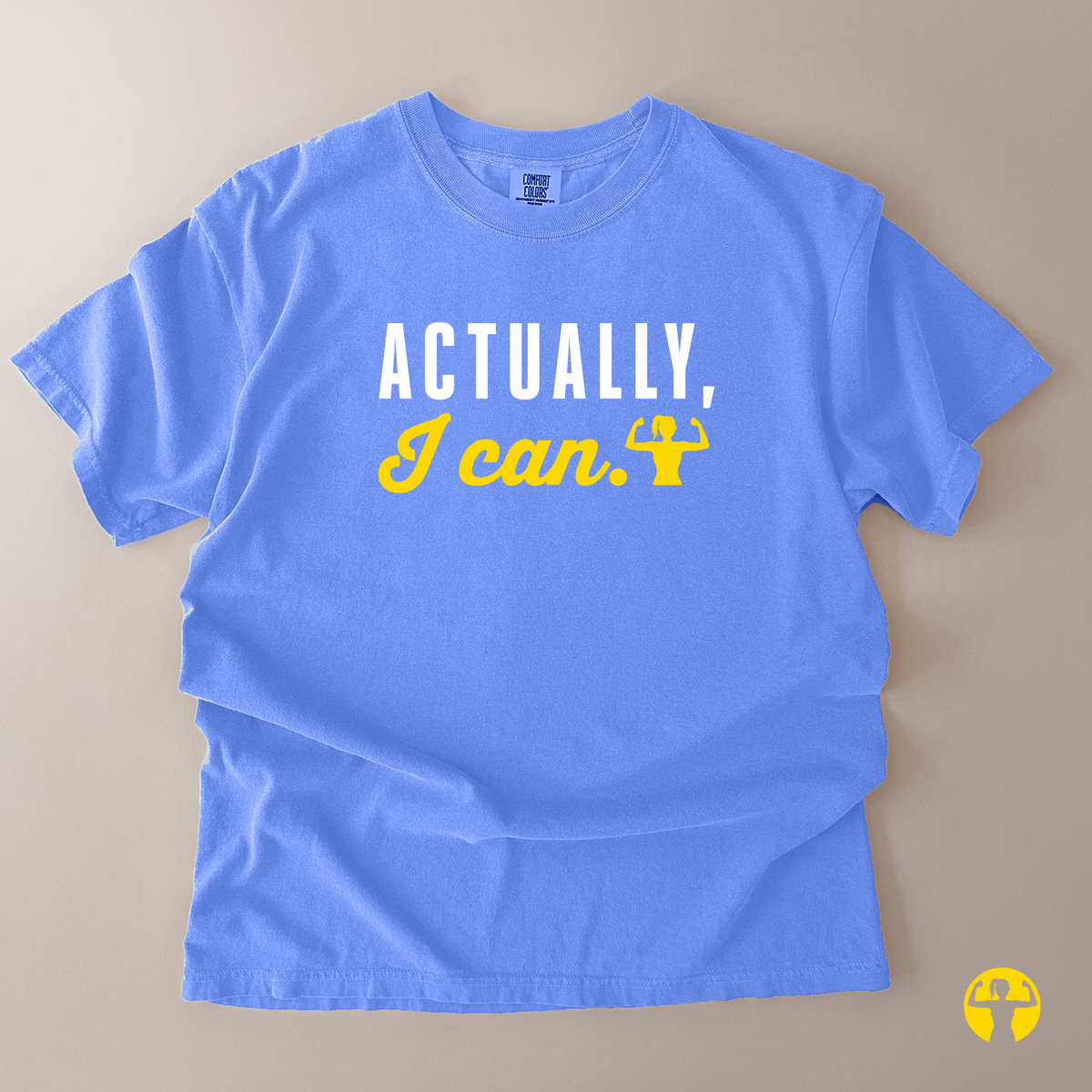 A light blue short-sleeve t-shirt with the text 'ACTUALLY, I can.' printed on the front in white and yellow, laid flat on a beige background.