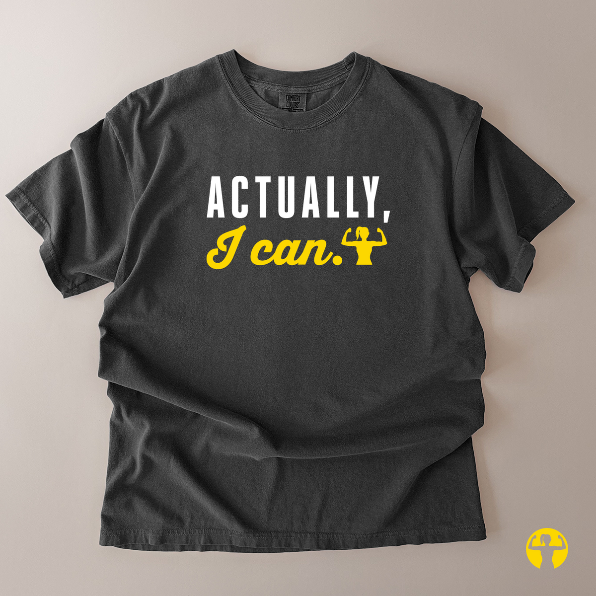 A dark charcoal gray t-shirt laid flat on a light beige background, featuring the text 'ACTUALLY, I can.' in white and yellow with a small yellow flexing bicep graphic.