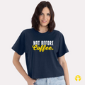 A smiling woman with dark, shoulder-length hair models a navy blue crewneck crop tee. The front of the shirt features a graphic text design that reads "NOT BEFORE COFFEE" with "NOT BEFORE" printed in bold white block letters and "Coffee" written in bright yellow cursive script. The background is solid light gray, and a yellow circular logo featuring a strong woman symbol is visible in the bottom right corner of the image.
