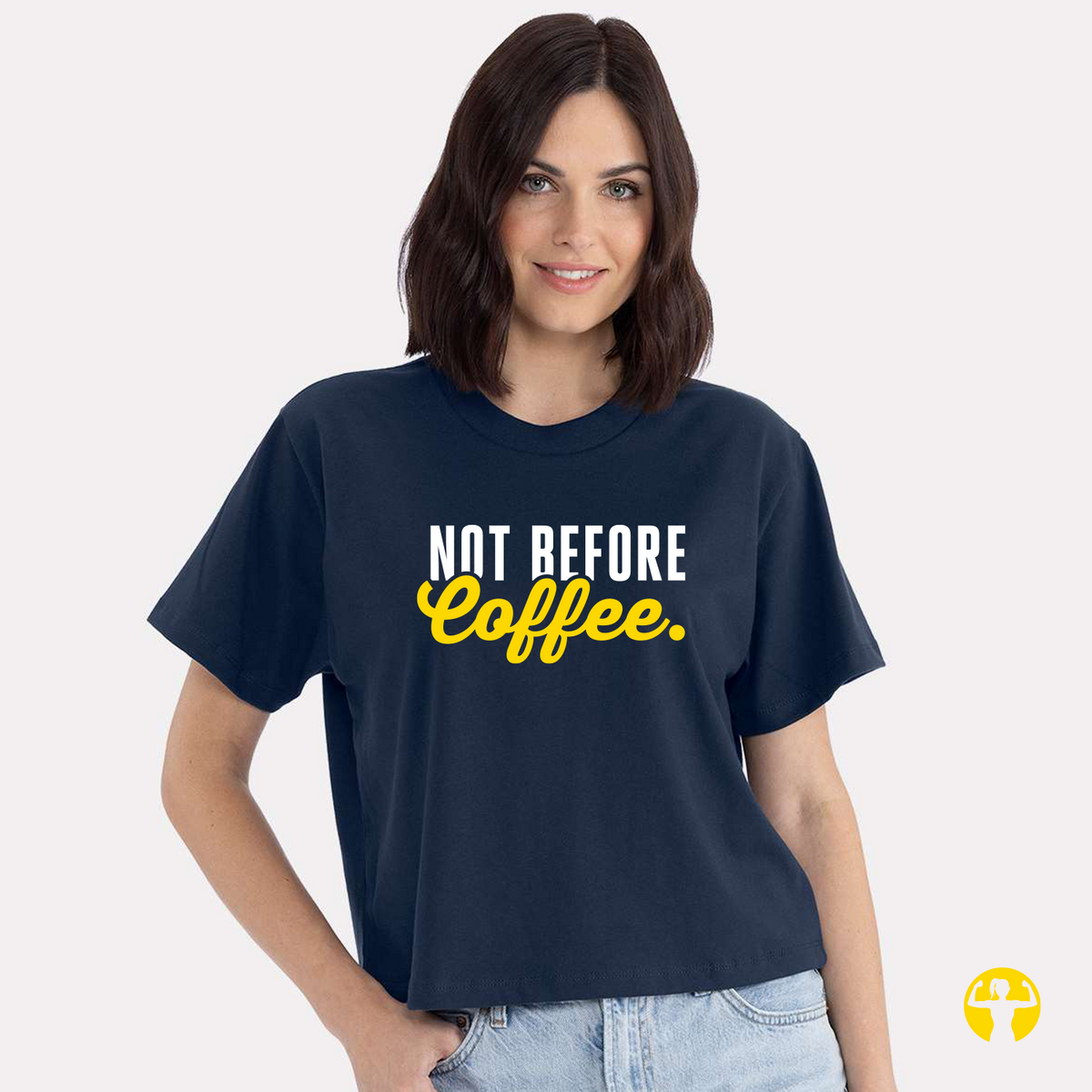 A smiling woman with dark, shoulder-length hair models a navy blue crewneck crop tee. The front of the shirt features a graphic text design that reads "NOT BEFORE COFFEE" with "NOT BEFORE" printed in bold white block letters and "Coffee" written in bright yellow cursive script. The background is solid light gray, and a yellow circular logo featuring a strong woman symbol is visible in the bottom right corner of the image.