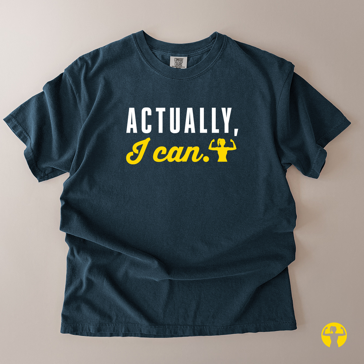 A dark navy blue t-shirt laid flat on a light beige background, featuring the text 'ACTUALLY, I can.' printed on the chest in white and yellow, with a small yellow flexing arm graphic.