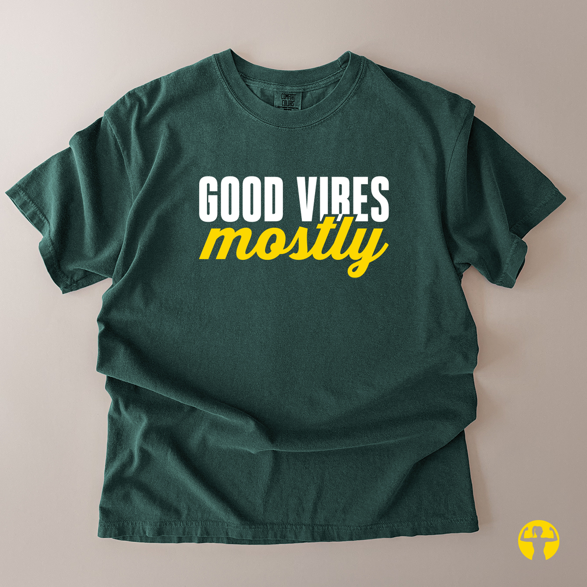 A dark green short-sleeve t-shirt laid flat on a light beige background, featuring the text 'GOOD VIBES mostly' printed on the chest in white and yellow.