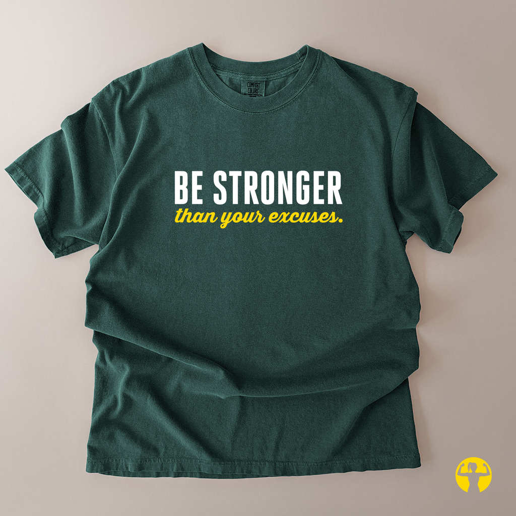 A dark green short-sleeve t-shirt laid flat on a light beige background, featuring the text 'BE STRONGER' in white and 'than your excuses.' in yellow across the chest.
