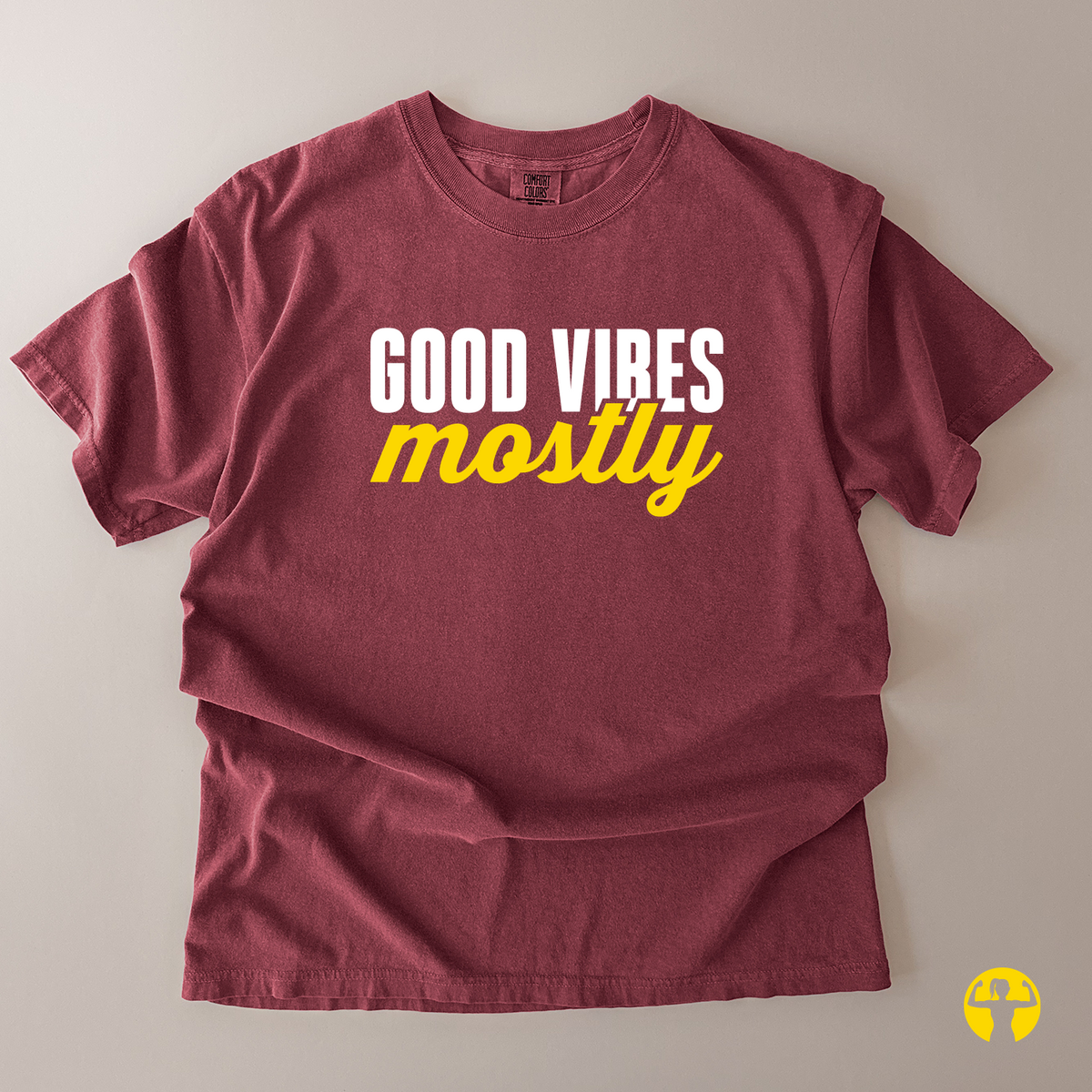 A maroon short-sleeve t-shirt with 'GOOD VIBES mostly' printed on the chest, laid flat on a neutral beige background. The text'GOOD VIBES' is in white block letters, and 'mostly' is in yellow script.