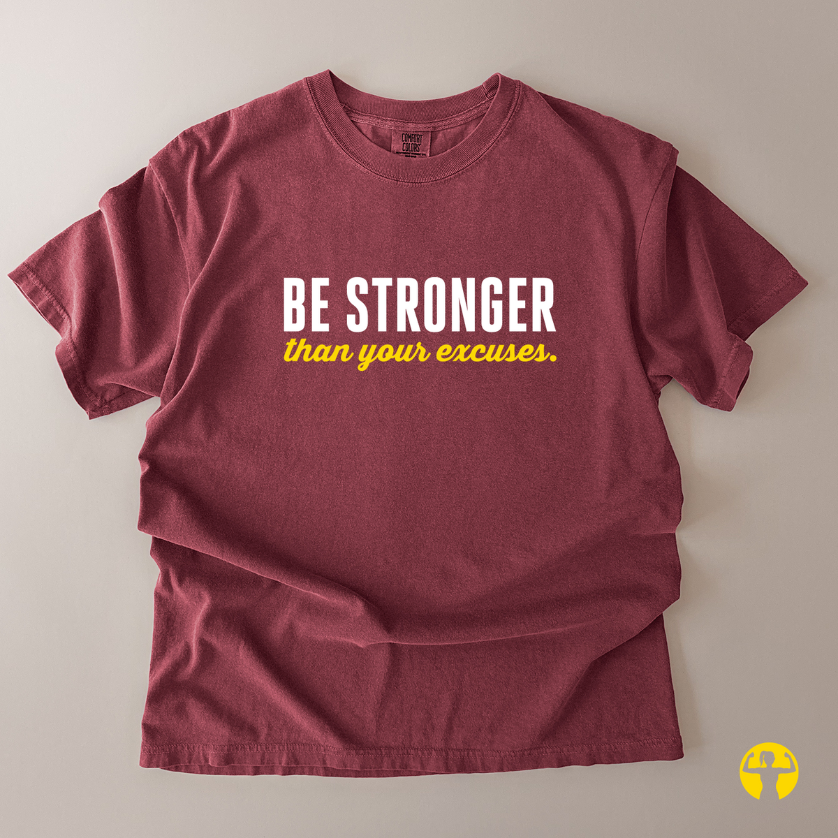 A maroon short-sleeve t-shirt with the text 'BE STRONGER than your excuses.' printed on the chest, laid flat on a neutral beige background.