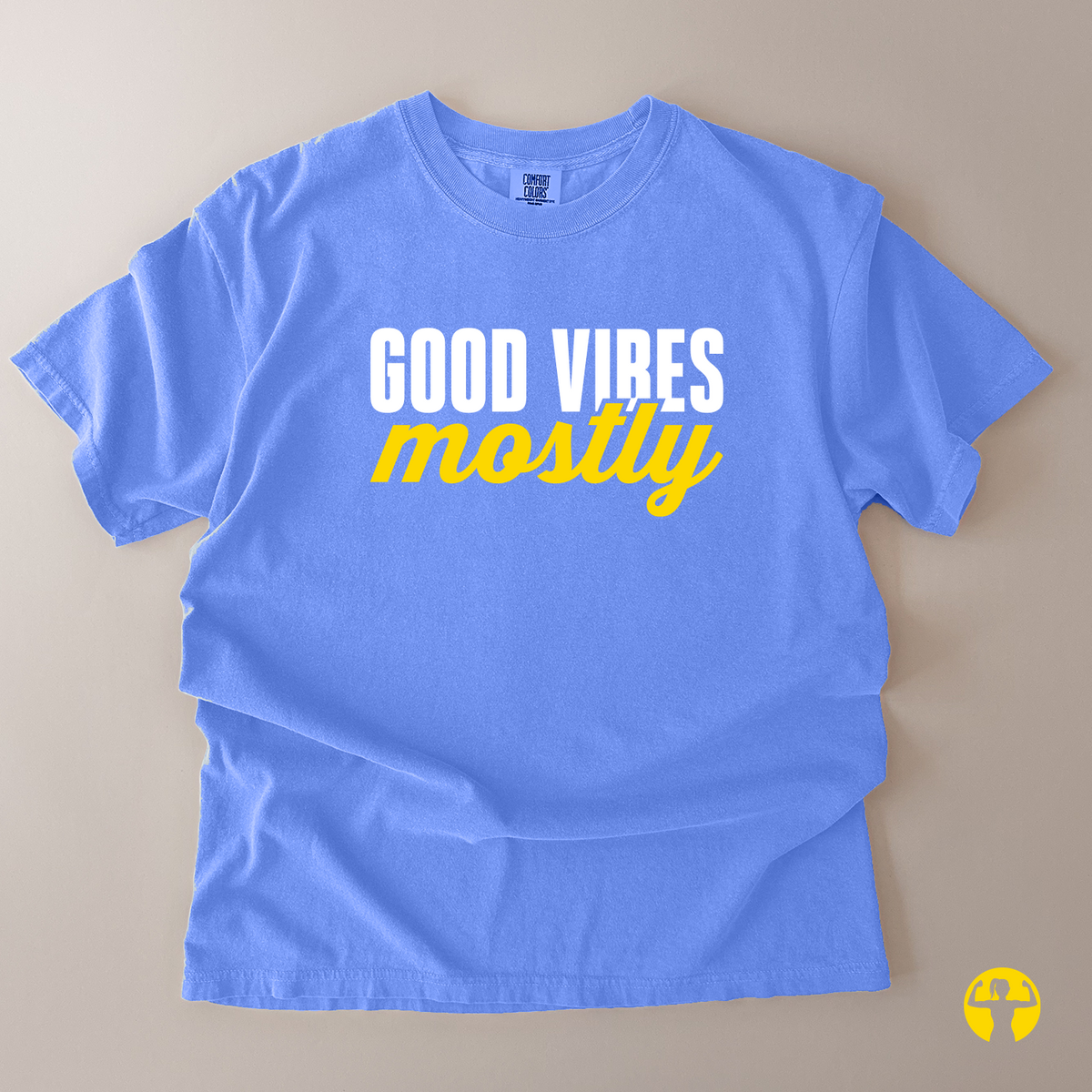 A light blue short-sleeve t-shirt laid flat on a beige background, featuring the text 'GOOD VIRES mostly' printed on the chest in white and yellow.