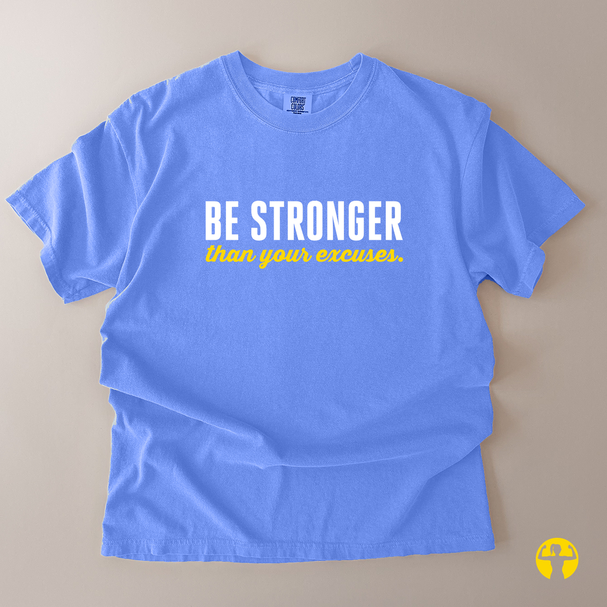 A light blue short-sleeve t-shirt laid flat on a beige background, featuring the text 'BE STRONGER' in white block letters and 'than your excuses.' in yellow script below it.