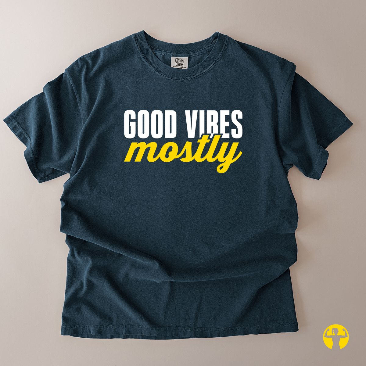 A dark navy blue t-shirt laid flat on a light beige background, featuring the text 'GOOD VIBES mostly' printed on the chest in white and yellow.