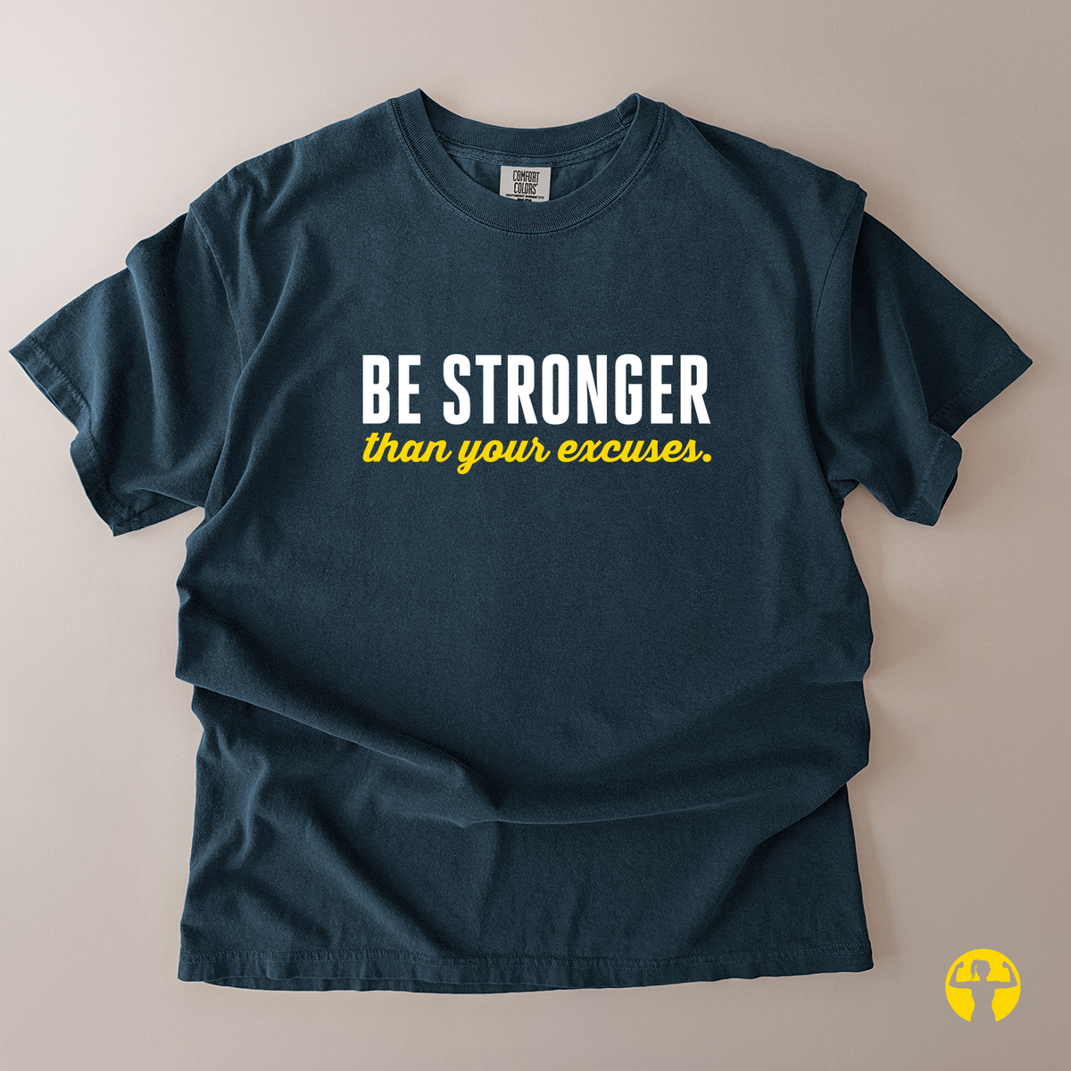 A dark navy blue t-shirt laid flat on a light beige background, featuring the text 'BE STRONGER than your excuses.' printed on the chest in white and yellow.