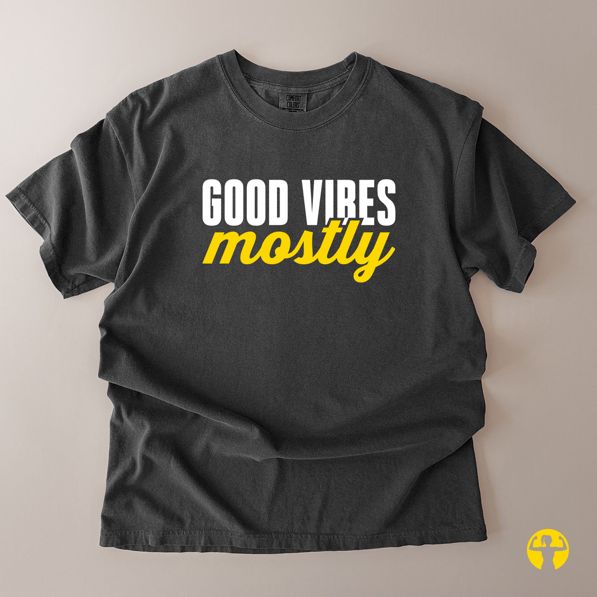 A dark charcoal gray t-shirt laid flat on a neutral beige background, featuring the text 'GOOD VIBES mostly' printed on the chest in white and yellow.