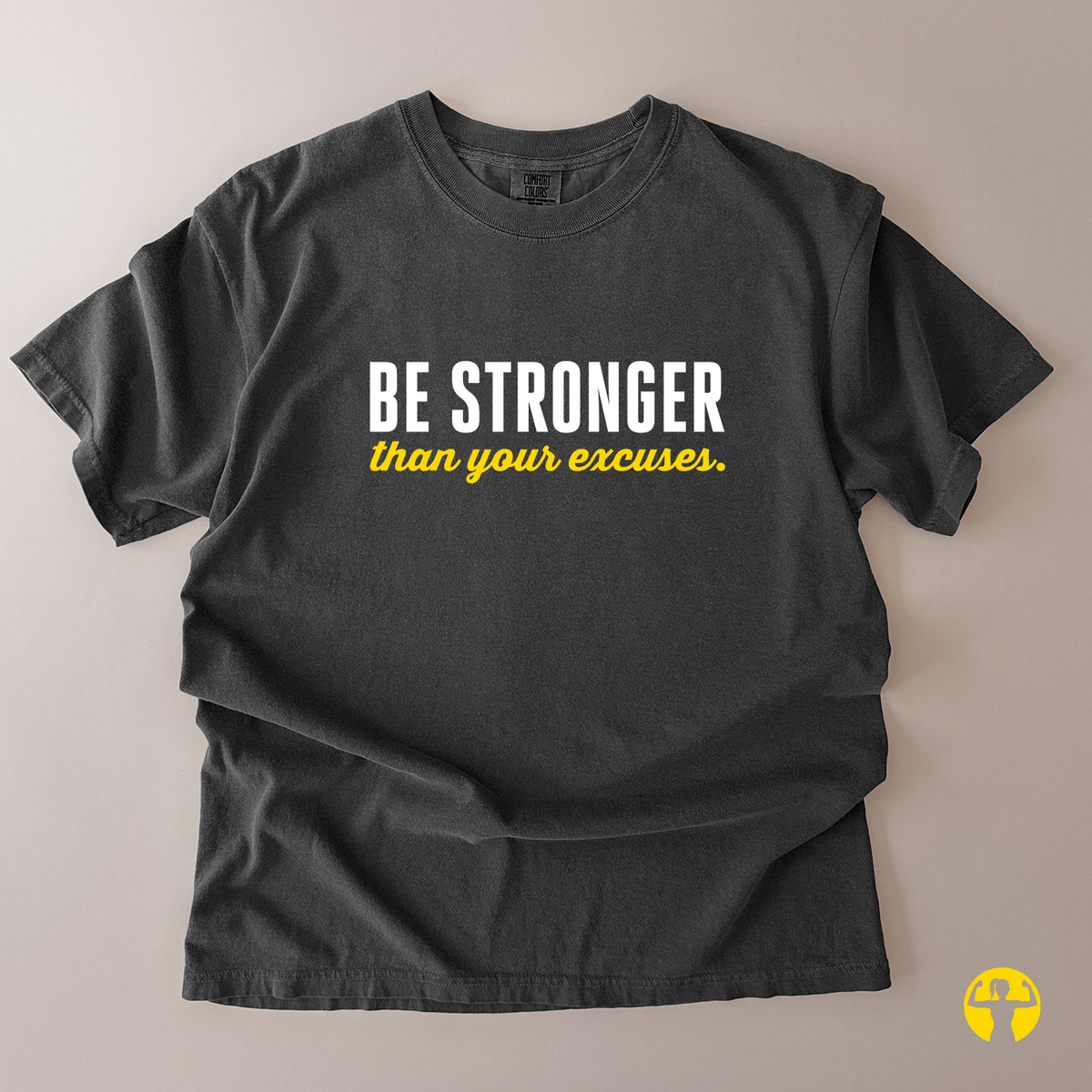 A dark charcoal gray t-shirt with the motivational quote 'BE STRONGER than your excuses.' printed on the chest in white and yellow, laid flat on a neutral beige background.