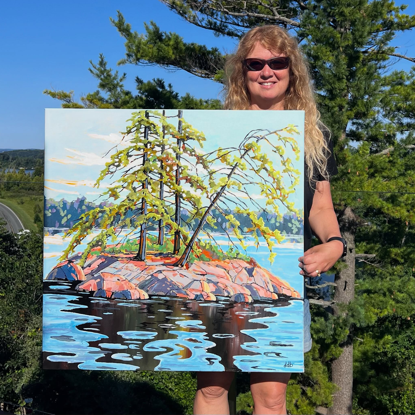 A woman wearing sunglasses holds up a colorful painting of a small rocky island with trees reflected in blue water, photographed outdoors in front of trees and a blue sky.