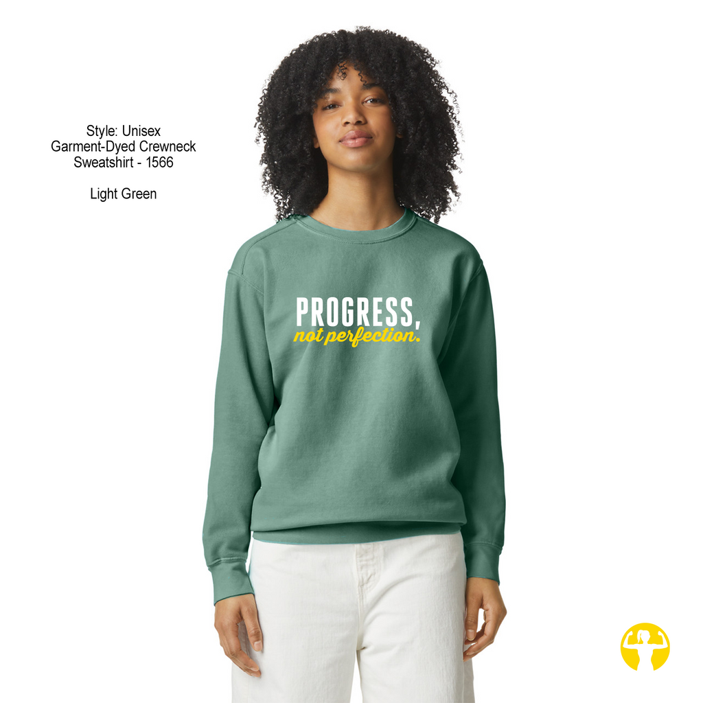 A model wearing a light green crewneck sweatshirt with the text 'PROGRESS, not perfection.' printed on the chest in white and yellow.