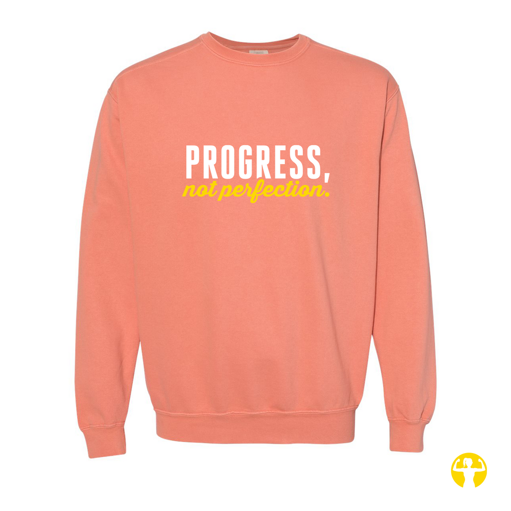 A coral pink crewneck sweatshirt featuring the text 'PROGRESS, not perfection' printed on the chest in white and yellow, displayed against a solid white background.