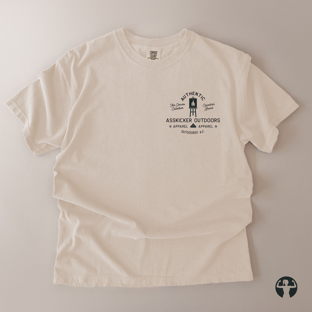 An off-white short-sleeve t-shirt with a black graphic print on the left chest, laid flat on a neutral beige background. The graphic features a painter's canval illustration and the text 'AUTHENTIC' and 'ASSKICKER OUTDOORS' 