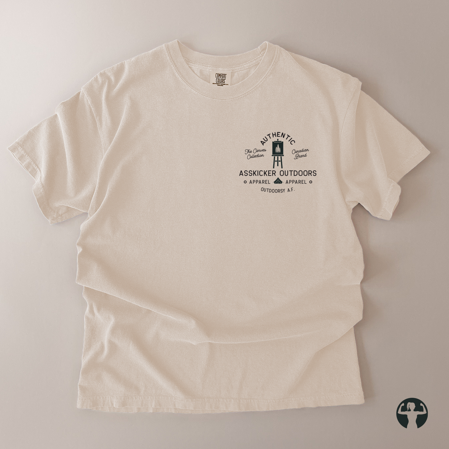An off-white short-sleeve t-shirt with a black graphic print on the left chest, laid flat on a neutral beige background. The graphic features a painter's canval illustration and the text 'AUTHENTIC' and 'ASSKICKER OUTDOORS' 
