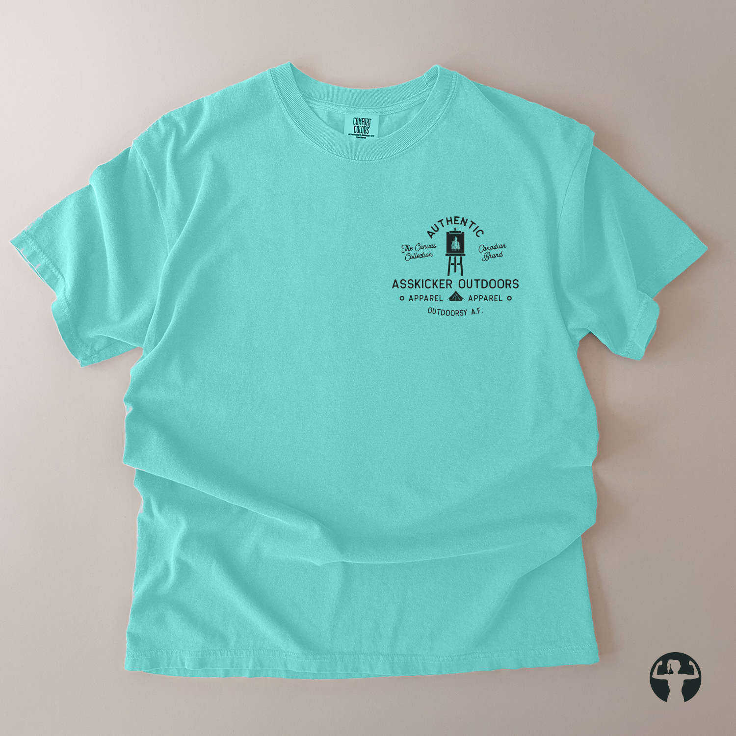 A light turquoise crewneck t-shirt laid flat on a neutral beige background. The shirt features a black graphic print on the left chest with the text 'AUTHENTIC ASSKICKER OUTDOORS' and a small illustration of a artist's easel.
