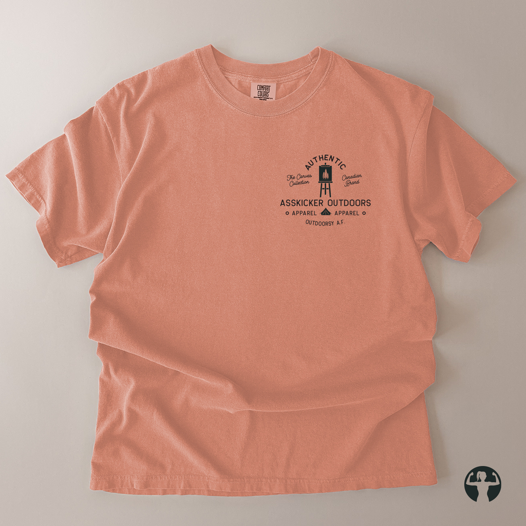 A peach-colored short-sleeve t-shirt with a black graphic print on the left chest, laid flat on a neutral gray background. The graphic features a lighthouse illustration and the text 'Authentic Asskicker Outdoors Appaarel'. A Canadian Brand.