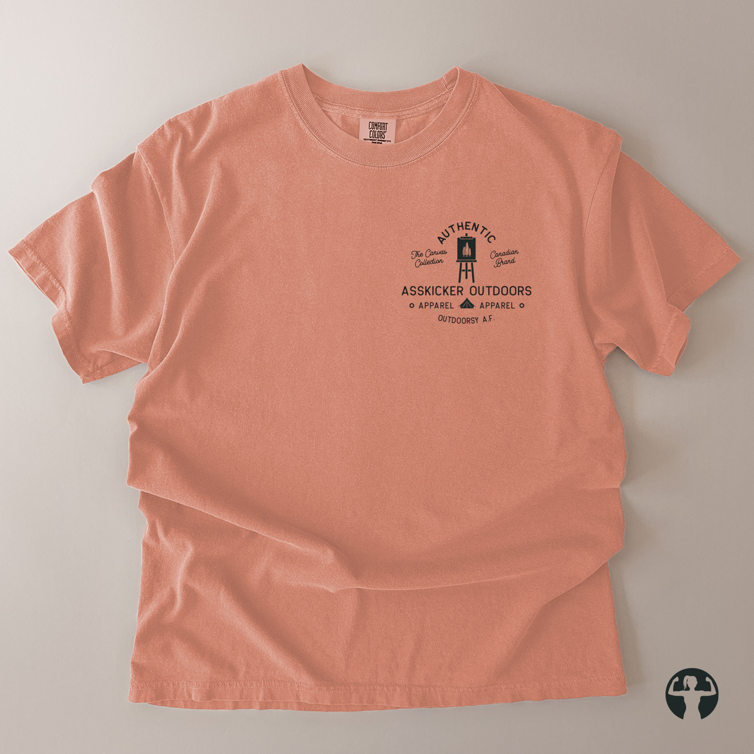 A peach-colored short-sleeve t-shirt with a black graphic print on the left chest, laid flat on a neutral gray background. The graphic features a lighthouse illustration and the text 'Authentic Asskicker Outdoors Appaarel'. A Canadian Brand.