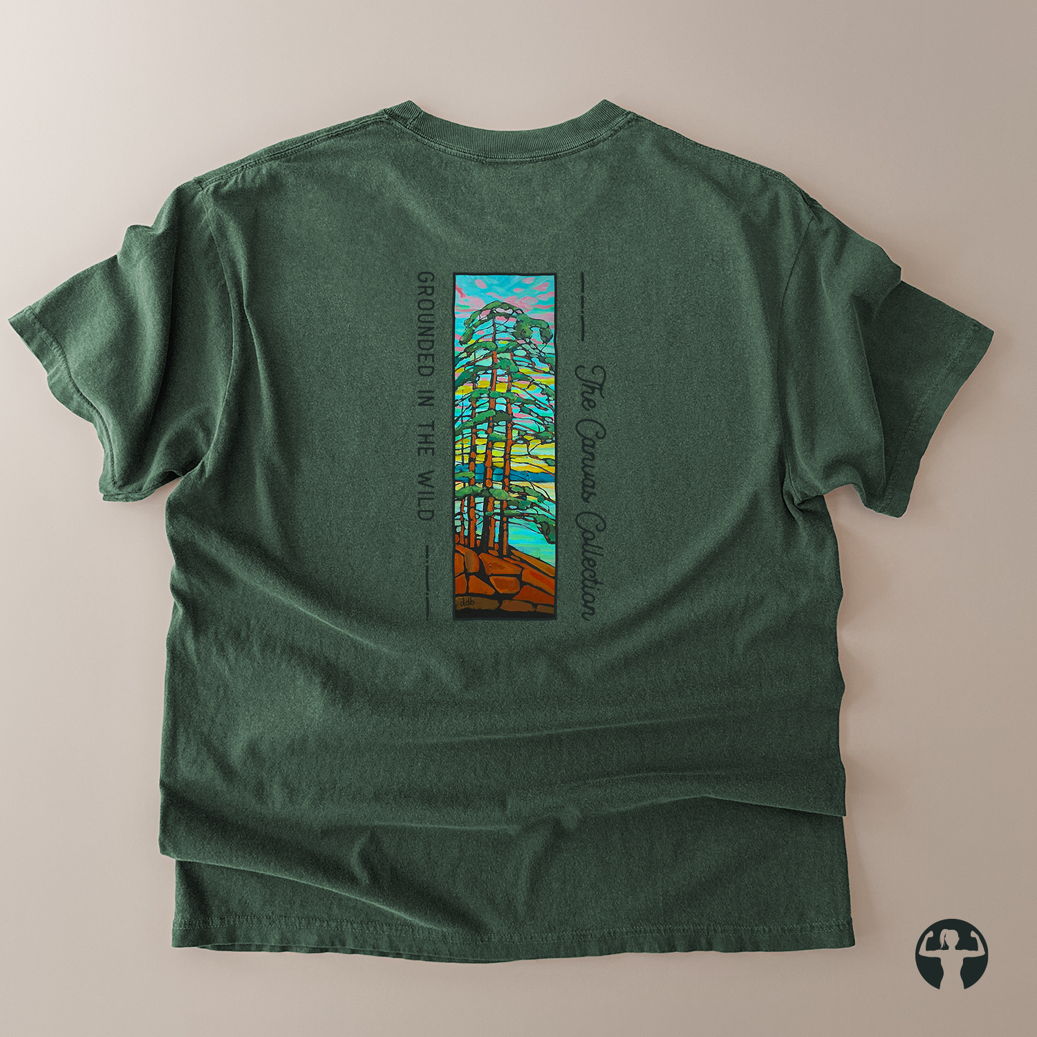A dark green short-sleeve t-shirt laid flat on a light beige background, showing the back side with a colorful graphic of pine trees and text reading 'GROUNDED IN THE WILD' and 'The Canvas Collection'.