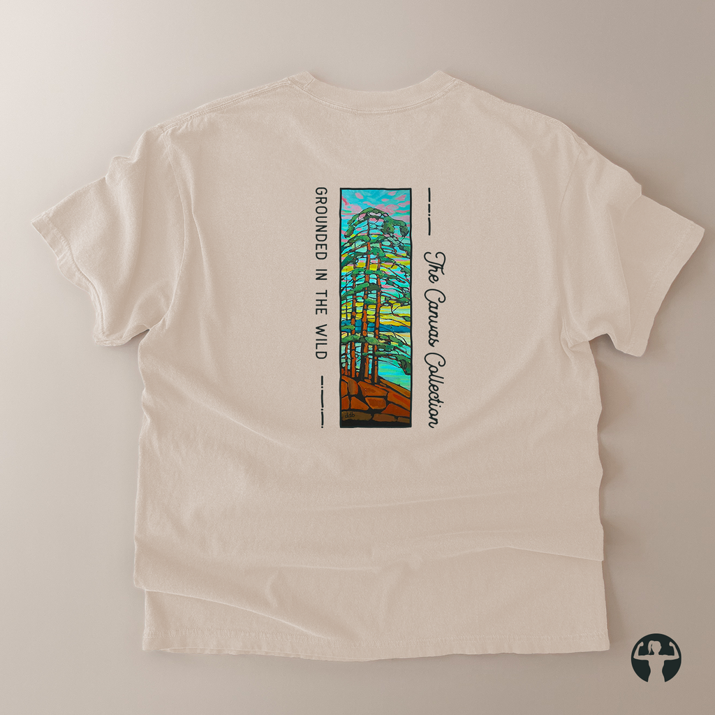 An off-white short-sleeve t-shirt laid flat on a neutral beige background, showing the back side with a colorful graphic of pine trees and the text 'GROUNDED IN THE WILD' and 'The Canvas Collection'.